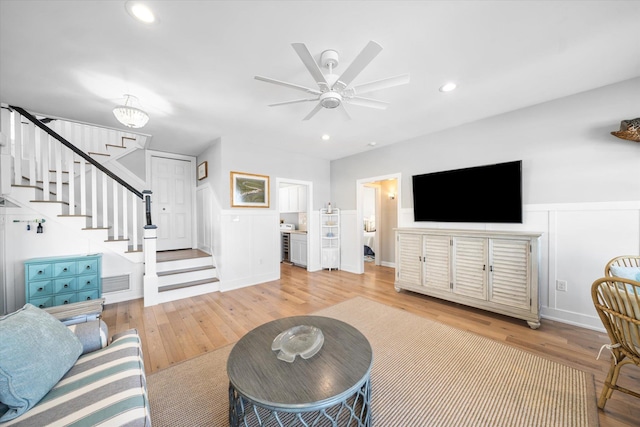 living area featuring stairway, wainscoting, ceiling fan, a decorative wall, and light wood-style floors