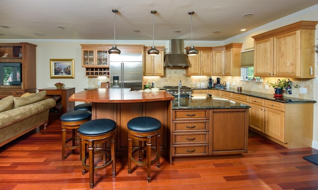 kitchen featuring built in fridge, wall chimney exhaust hood, an island with sink, and decorative light fixtures