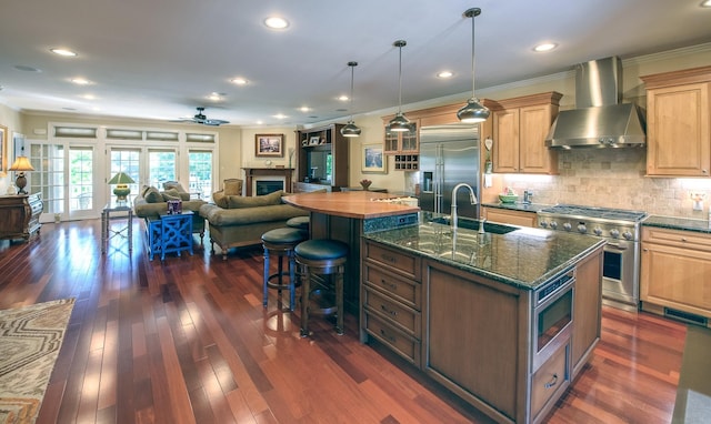 kitchen featuring ceiling fan, sink, wall chimney range hood, built in appliances, and a center island with sink