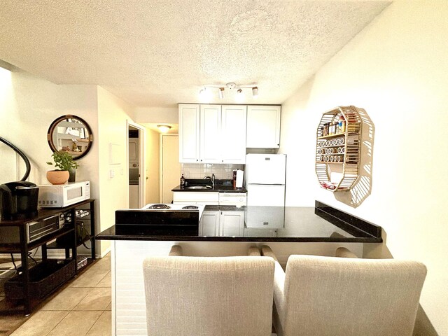 kitchen featuring a kitchen bar, white appliances, a textured ceiling, light tile patterned floors, and white cabinetry