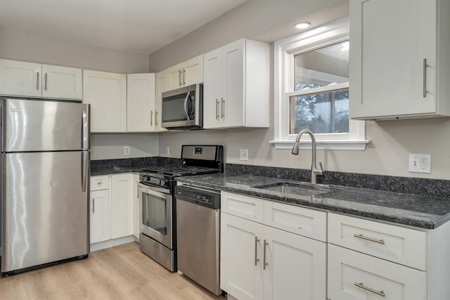 kitchen with appliances with stainless steel finishes, sink, white cabinets, dark stone counters, and light hardwood / wood-style floors