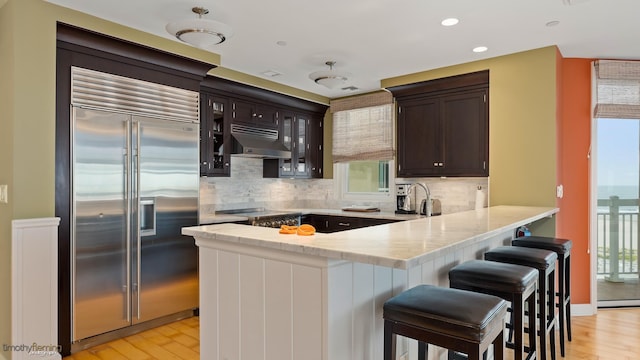 kitchen with under cabinet range hood, a peninsula, light wood-style floors, dark brown cabinets, and stainless steel built in fridge