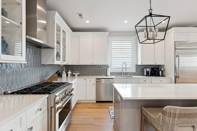 kitchen with wall chimney exhaust hood, high end appliances, white cabinetry, and hanging light fixtures