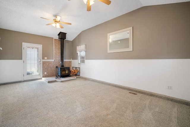 unfurnished living room with a wainscoted wall, vaulted ceiling, a ceiling fan, a wood stove, and a textured ceiling