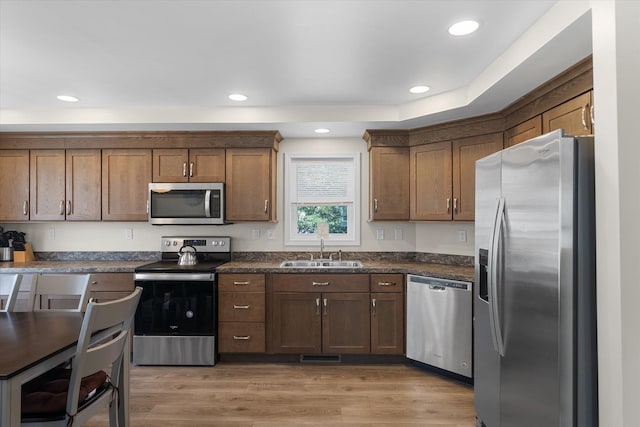 kitchen featuring appliances with stainless steel finishes, light wood-type flooring, a tray ceiling, sink, and dark stone countertops