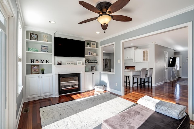 living area with dark wood-type flooring, visible vents, baseboards, a glass covered fireplace, and crown molding