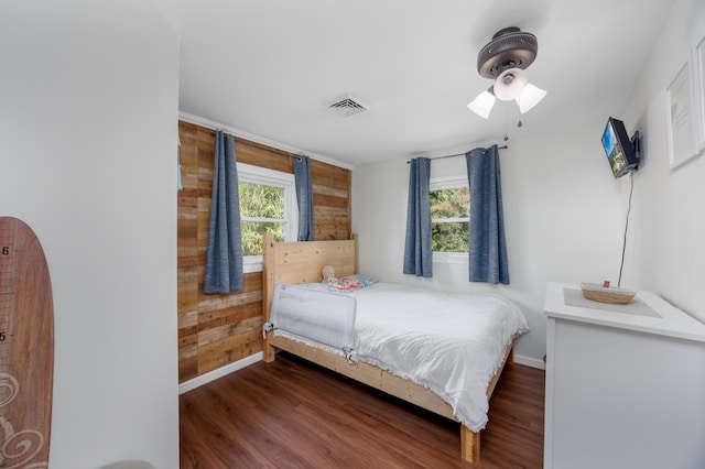 bedroom featuring ceiling fan, dark hardwood / wood-style flooring, wooden walls, and multiple windows