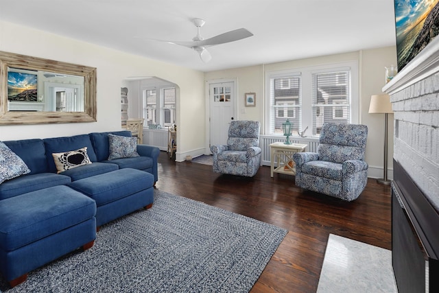living room featuring baseboards, arched walkways, a brick fireplace, ceiling fan, and wood finished floors