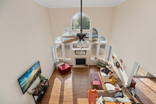 living room with a towering ceiling, a fireplace with flush hearth, ceiling fan, ornamental molding, and light wood-type flooring