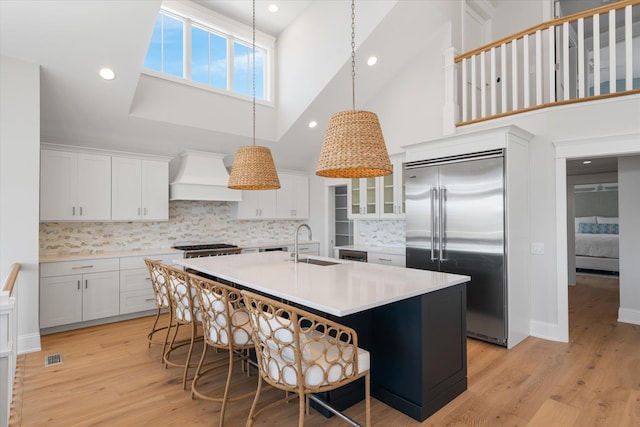 kitchen with built in fridge, premium range hood, a sink, light countertops, and a center island with sink