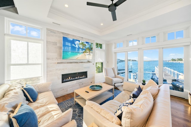 living room with dark wood-type flooring, a raised ceiling, crown molding, a tiled fireplace, and a water view