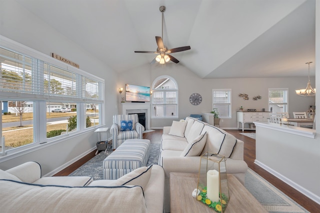 living room featuring baseboards, a fireplace with flush hearth, lofted ceiling, ceiling fan with notable chandelier, and dark wood-style floors