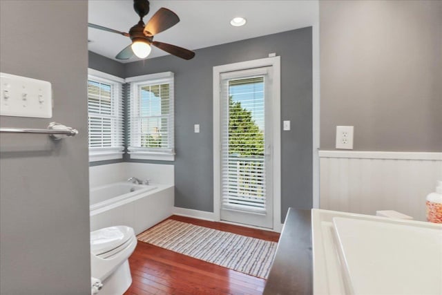 full bathroom with ceiling fan, a garden tub, toilet, a wainscoted wall, and wood-type flooring