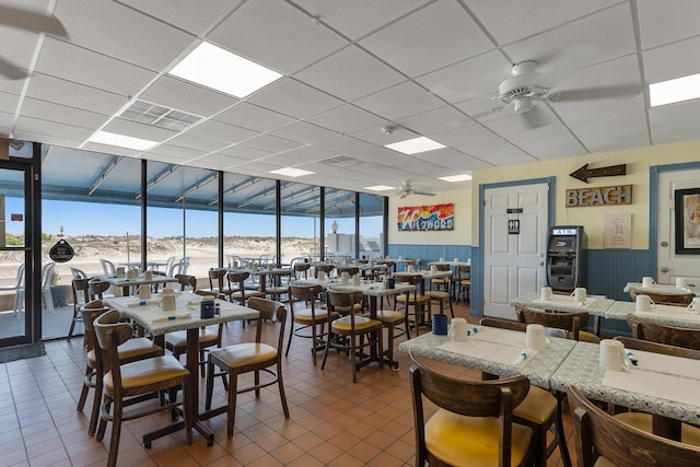 dining space with a ceiling fan, floor to ceiling windows, a drop ceiling, dark tile patterned floors, and a wainscoted wall