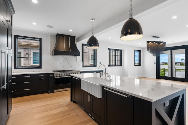 kitchen with light wood-type flooring, custom range hood, pendant lighting, a center island with sink, and stainless steel stove