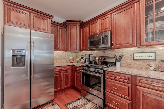 kitchen featuring light stone countertops, appliances with stainless steel finishes, light wood-type flooring, decorative backsplash, and glass insert cabinets