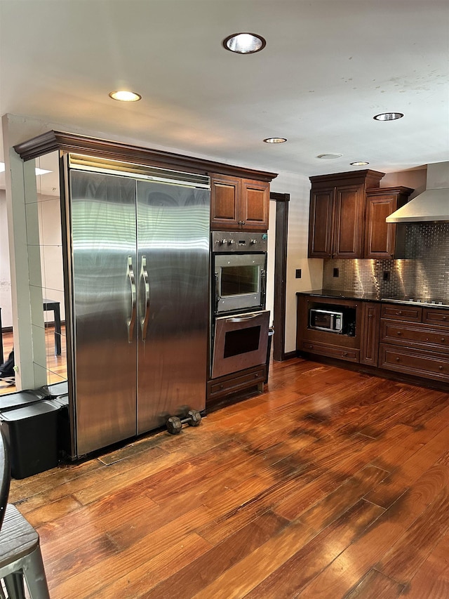 kitchen featuring wall chimney exhaust hood, dark wood-type flooring, double oven, built in refrigerator, and decorative backsplash