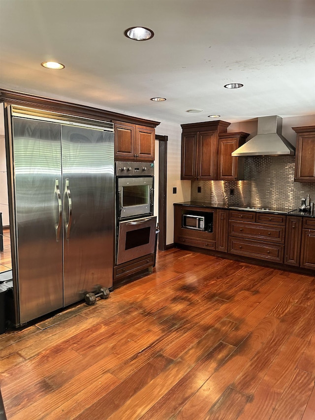 kitchen with wall chimney exhaust hood, dark hardwood / wood-style floors, stainless steel built in refrigerator, and multiple ovens