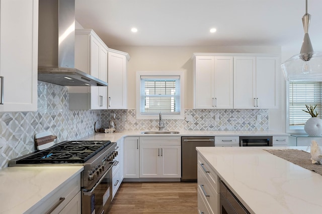 kitchen featuring sink, white cabinetry, hanging light fixtures, appliances with stainless steel finishes, and wall chimney range hood