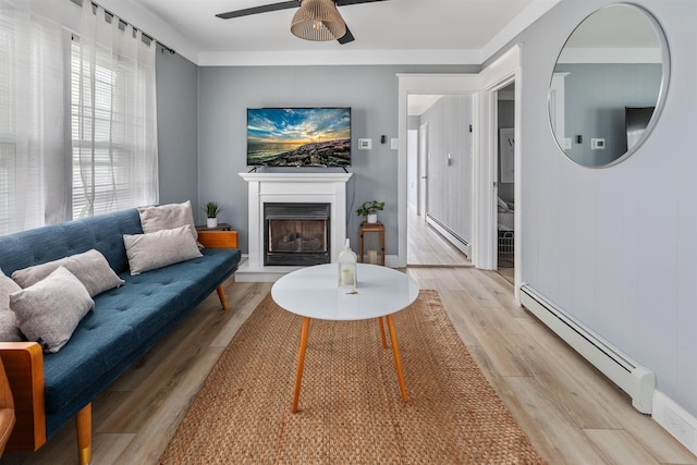 living room featuring a baseboard heating unit, a fireplace with raised hearth, a ceiling fan, and light wood-style floors