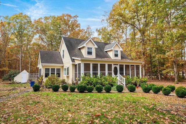 new england style home with a storage shed, a shingled roof, an outdoor structure, a sunroom, and a front yard