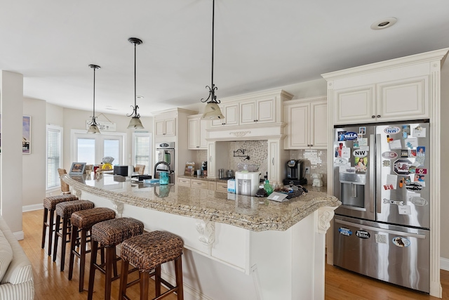 kitchen featuring stainless steel refrigerator with ice dispenser, hanging light fixtures, light stone countertops, an island with sink, and tasteful backsplash