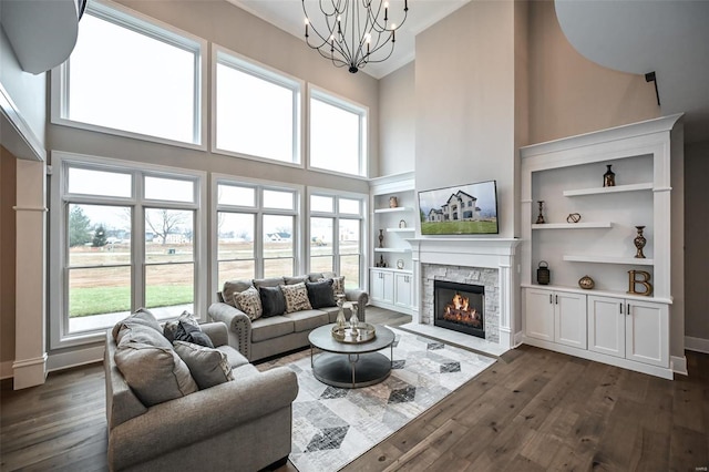 living room with dark wood-type flooring, a chandelier, a towering ceiling, built in shelves, and a stone fireplace