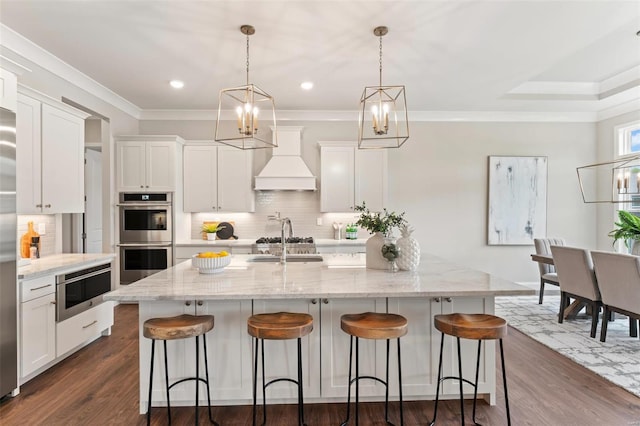 kitchen with double oven, decorative light fixtures, white cabinetry, a kitchen island with sink, and custom range hood