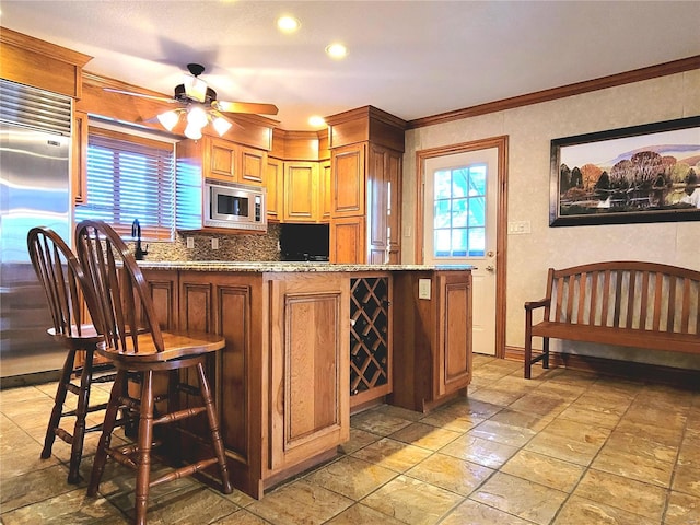 kitchen featuring ceiling fan, light stone countertops, a kitchen breakfast bar, built in appliances, and crown molding