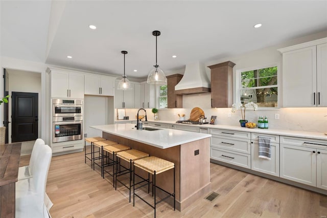 kitchen featuring premium range hood, appliances with stainless steel finishes, sink, white cabinets, and a center island with sink