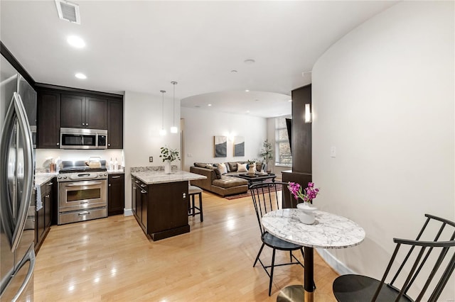 kitchen with hanging light fixtures, light stone counters, dark brown cabinets, appliances with stainless steel finishes, and light wood-type flooring