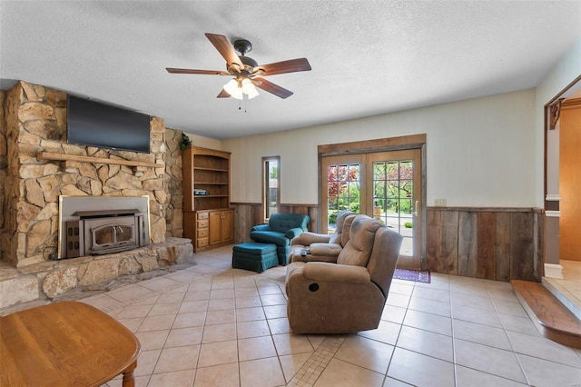 living room featuring wood walls, ceiling fan, a wood stove, light tile patterned floors, and a textured ceiling