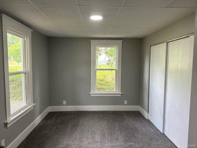 unfurnished bedroom with dark colored carpet, multiple windows, and a drop ceiling