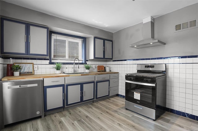 kitchen with wooden counters, appliances with stainless steel finishes, light wood-type flooring, sink, and wall chimney range hood