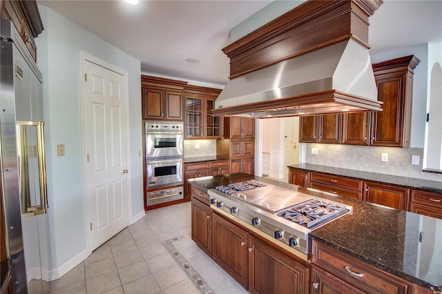 kitchen with dark stone counters, custom range hood, light tile flooring, stainless steel appliances, and tasteful backsplash