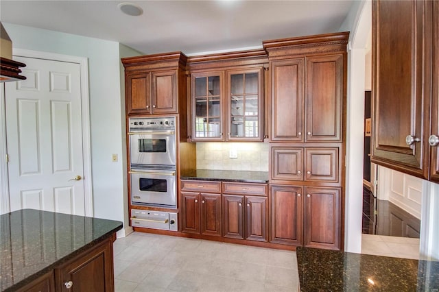 kitchen featuring stainless steel double oven, backsplash, light tile floors, and dark stone countertops