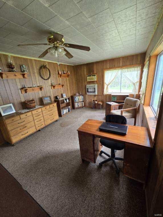 carpeted home office featuring wood walls and ceiling fan