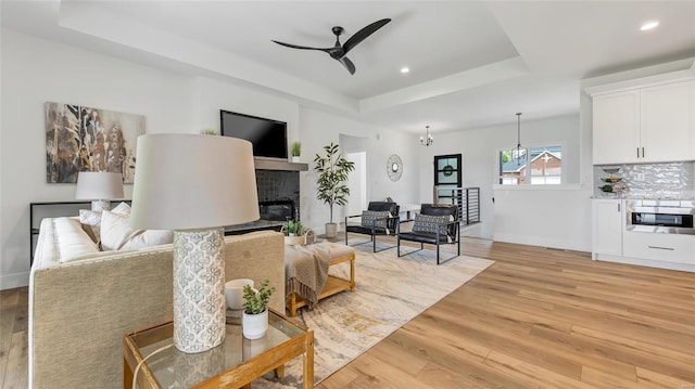 living room with a tray ceiling, a fireplace, ceiling fan with notable chandelier, and light wood-type flooring