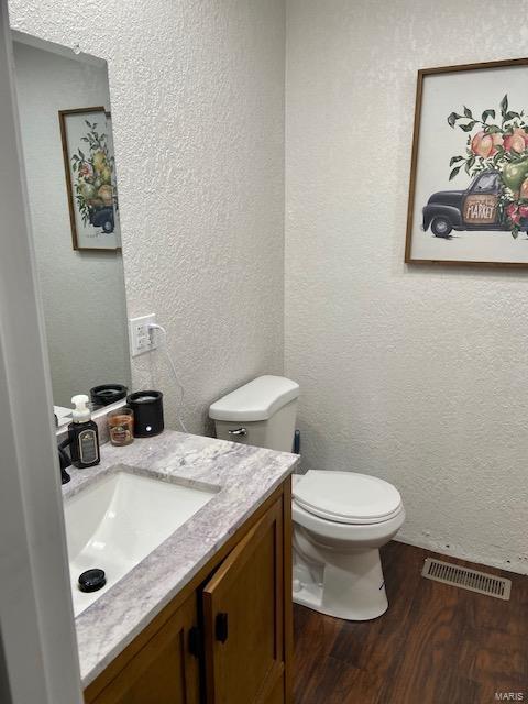 half bath featuring visible vents, a textured wall, toilet, wood finished floors, and vanity