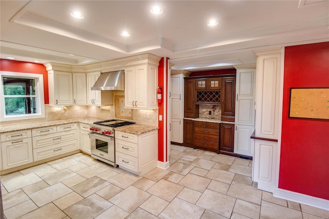 kitchen with wall chimney exhaust hood, light stone counters, a tray ceiling, designer stove, and decorative backsplash