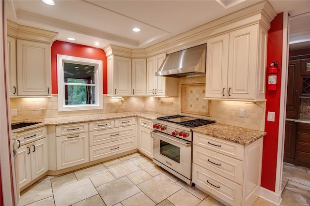 kitchen featuring backsplash, designer range, light stone counters, cream cabinets, and wall chimney range hood