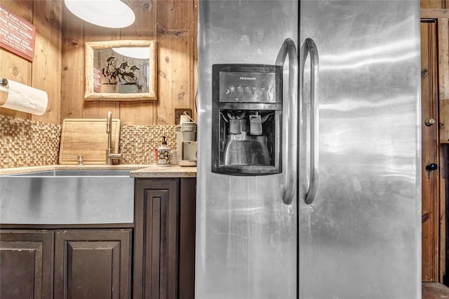 kitchen featuring backsplash, dark brown cabinetry, sink, stainless steel fridge with ice dispenser, and wood walls