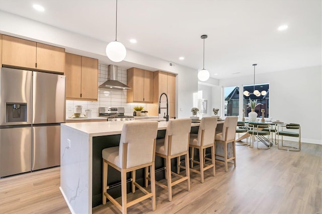 kitchen featuring a center island with sink, wall chimney exhaust hood, stainless steel appliances, and light brown cabinets