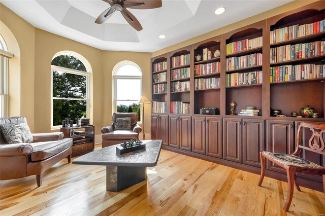 living area with ceiling fan, a raised ceiling, and light wood-type flooring