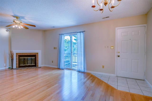 unfurnished living room with ceiling fan with notable chandelier, a tile fireplace, a textured ceiling, and light hardwood / wood-style flooring