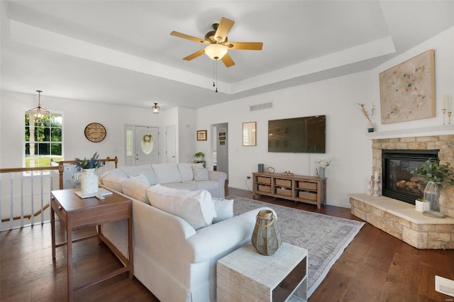 living room with dark wood-type flooring, ceiling fan, a tray ceiling, and a stone fireplace