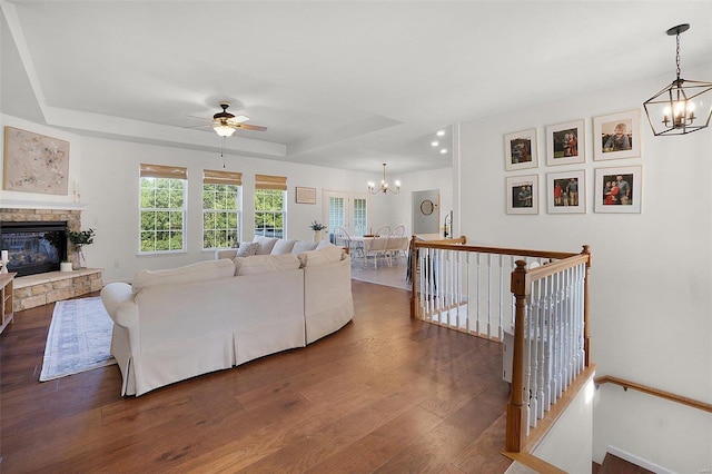 living room featuring a raised ceiling, ceiling fan with notable chandelier, dark hardwood / wood-style floors, and a stone fireplace