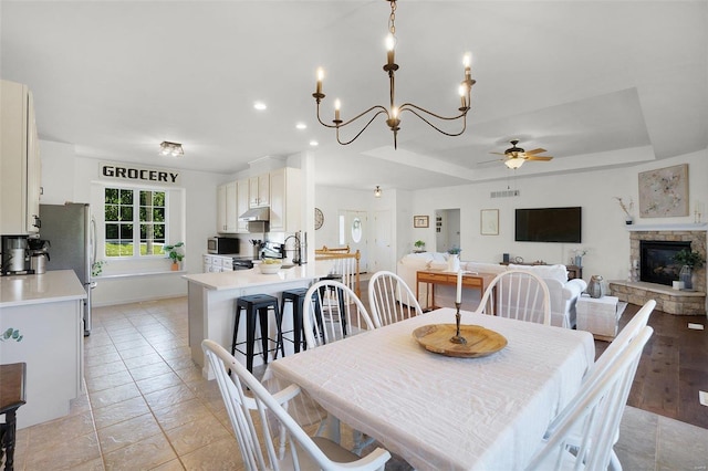 tiled dining space with a stone fireplace, sink, ceiling fan with notable chandelier, and a tray ceiling