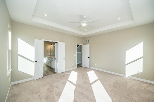 unfurnished bedroom featuring a tray ceiling, ensuite bath, ceiling fan, and light carpet