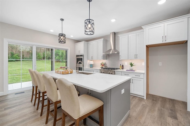 kitchen featuring a center island with sink, decorative backsplash, a sink, wall chimney range hood, and light wood-type flooring